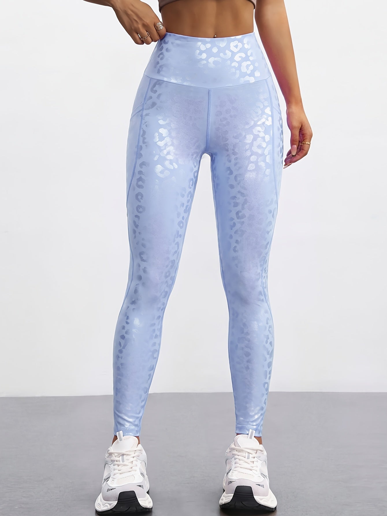 A model wearing high-waisted light blue leggings with a subtle white leopard print pattern, paired with white sneakers, shown from the waist down against a neutral studio background.